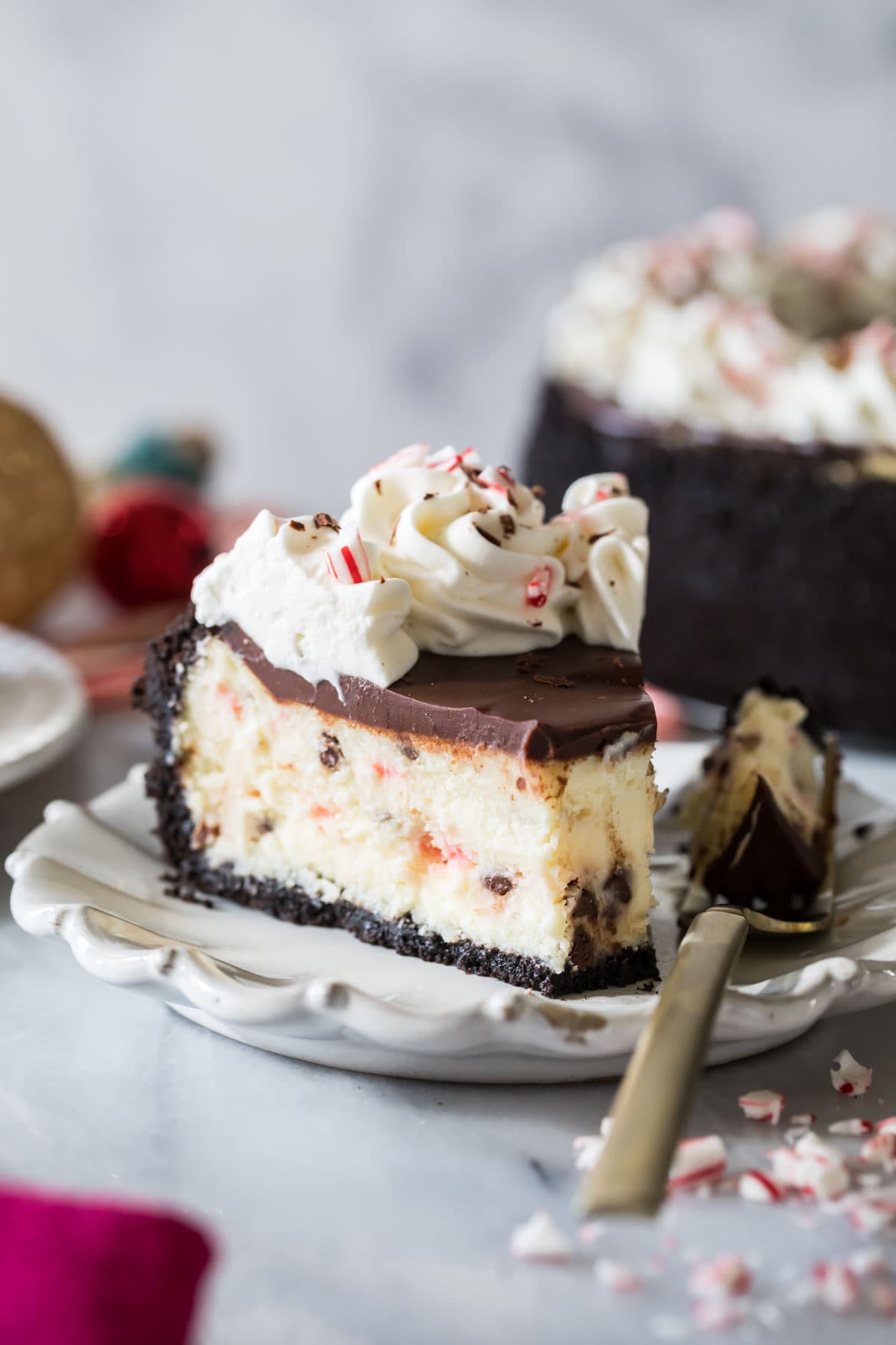 Slice of cheesecake topped with chocolate ganache, whipped cream, and crushed peppermint candy missing one bite.