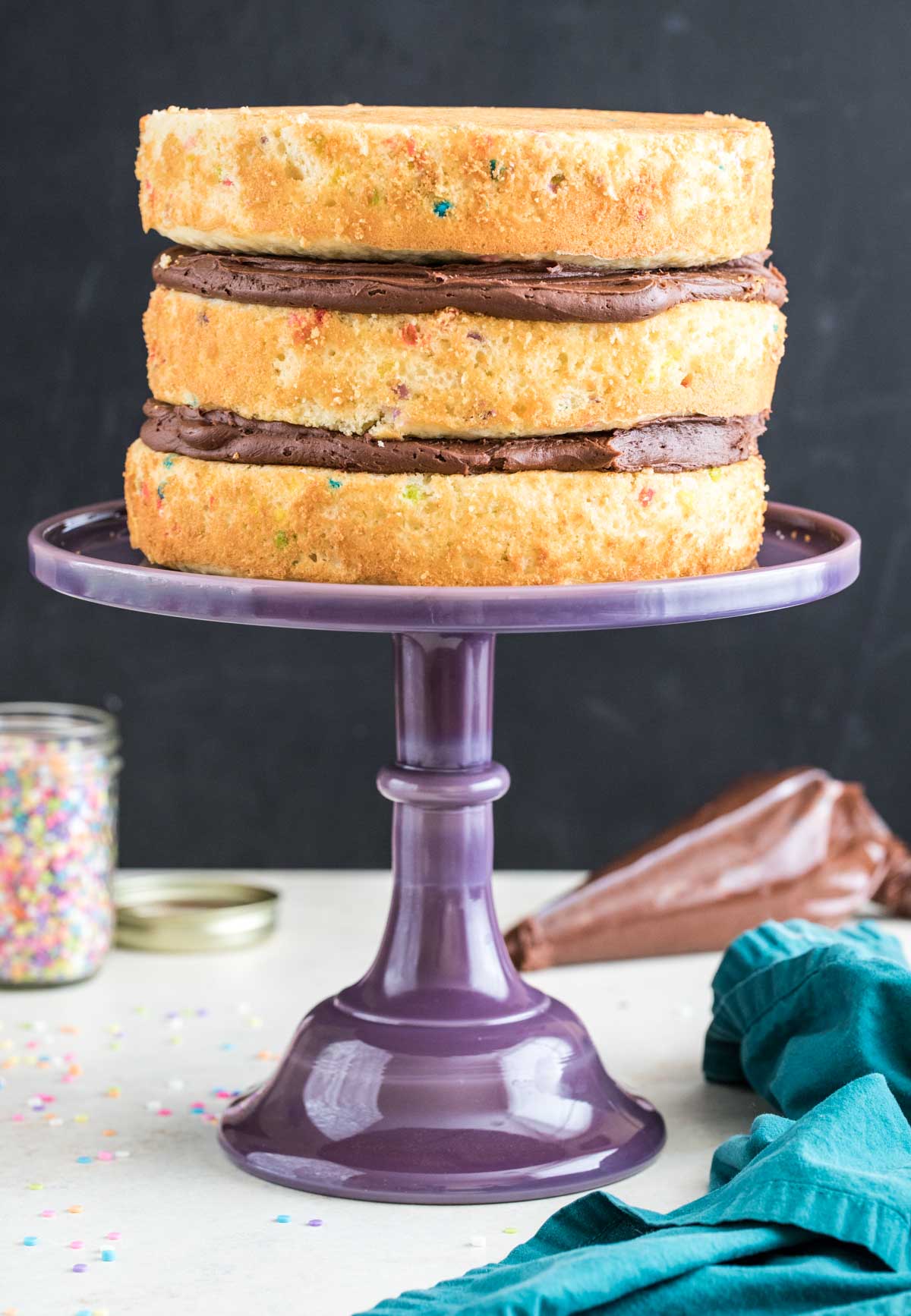 Purple cake stand with three layers of yellow cake stacked with chocolate frosting in between.