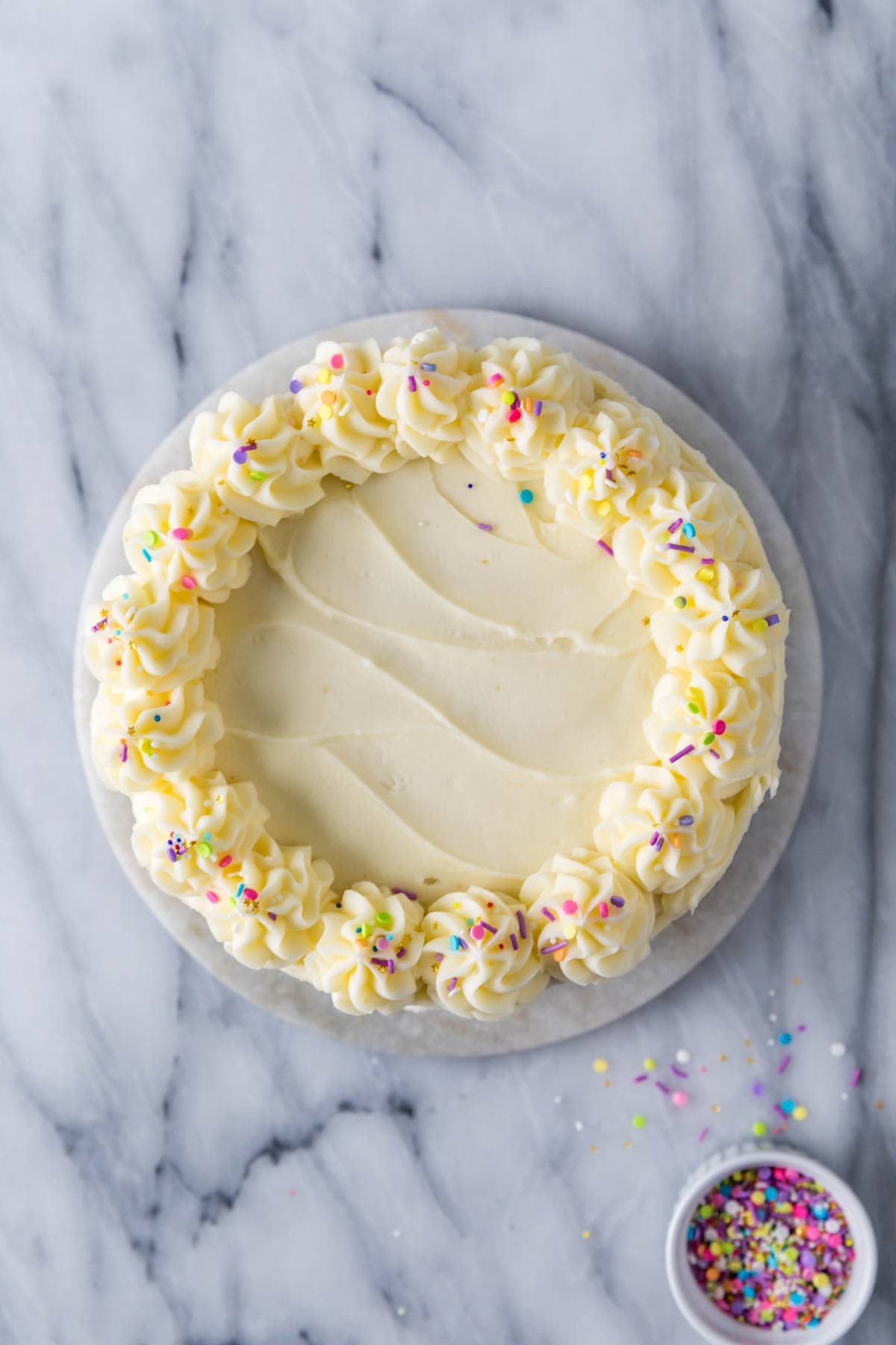 Overhead view of a cake frosted with white icing and a rim of piped icing swirls topped with sprinkles.