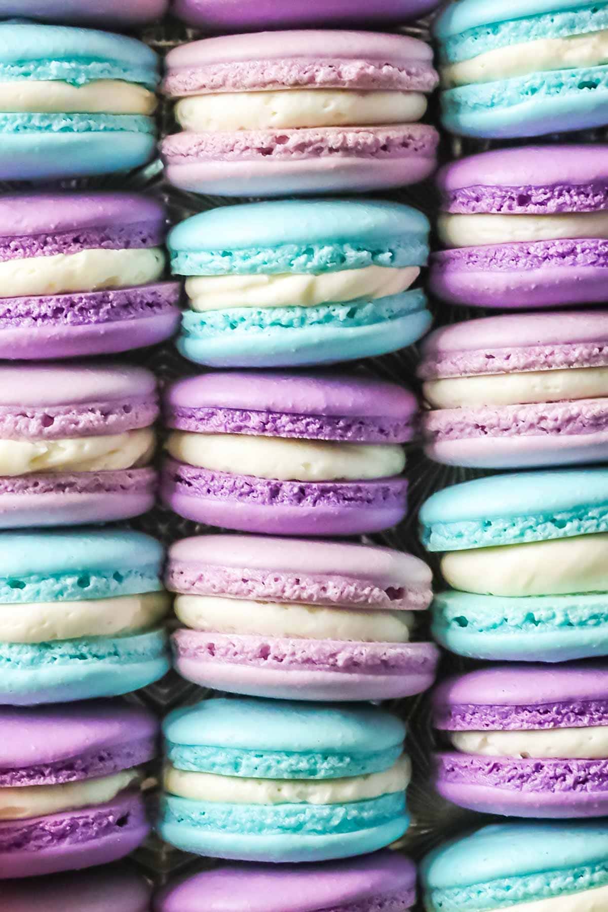 French macaron recipe showing side view of bright blue, light purple, and dark purple macarons sandwiches with white filling, all arranged in neat rows