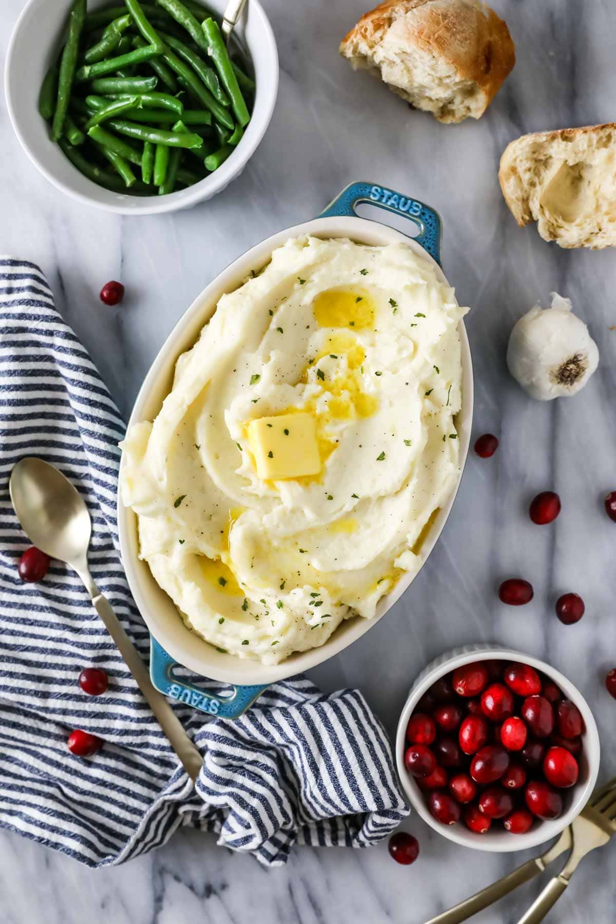 Overhead view of a casserole dish of mashed potatoes surrounded by green beans, cranberries, and bread.