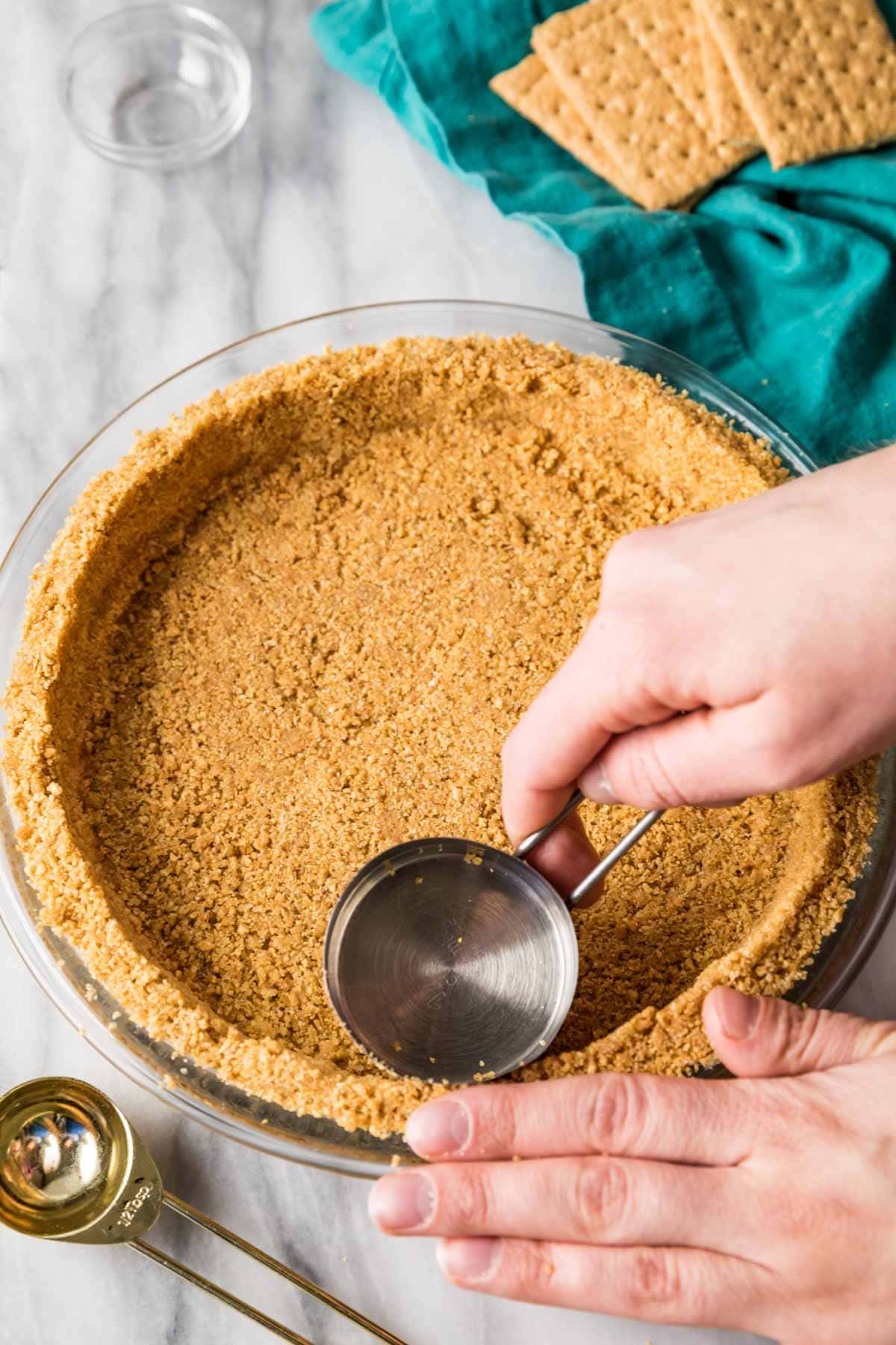 Using a metal measuring cup to tamp graham cracker crumbs into a glass pie plate.