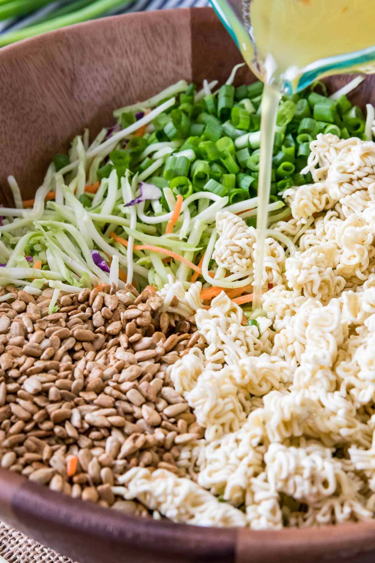 Dressing being poured onto a salad containing broccoli slaw, scallions, ramen noodles, and sunflower seeds.