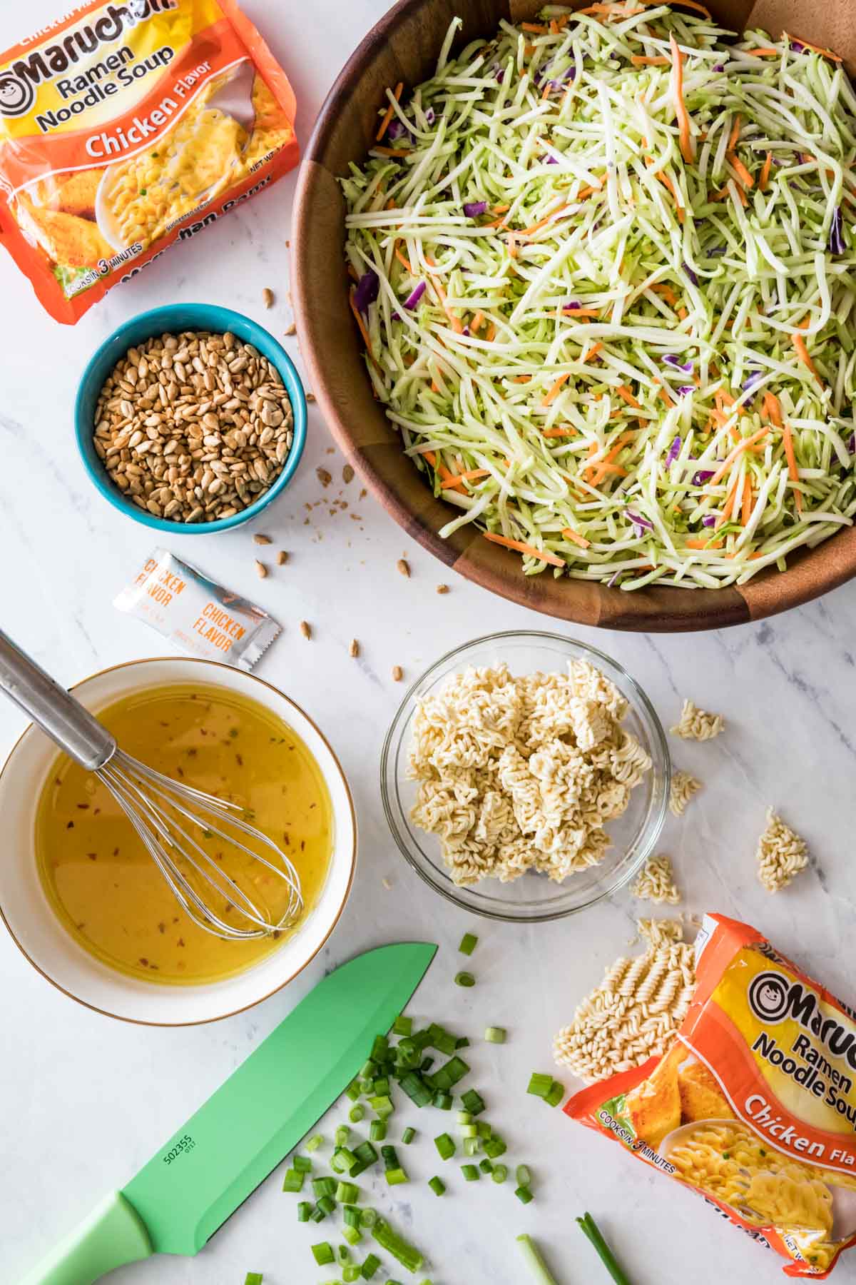 Overhead view of ingredients including broccoli slaw, ramen noodles, sunflower seeds, and scallions.