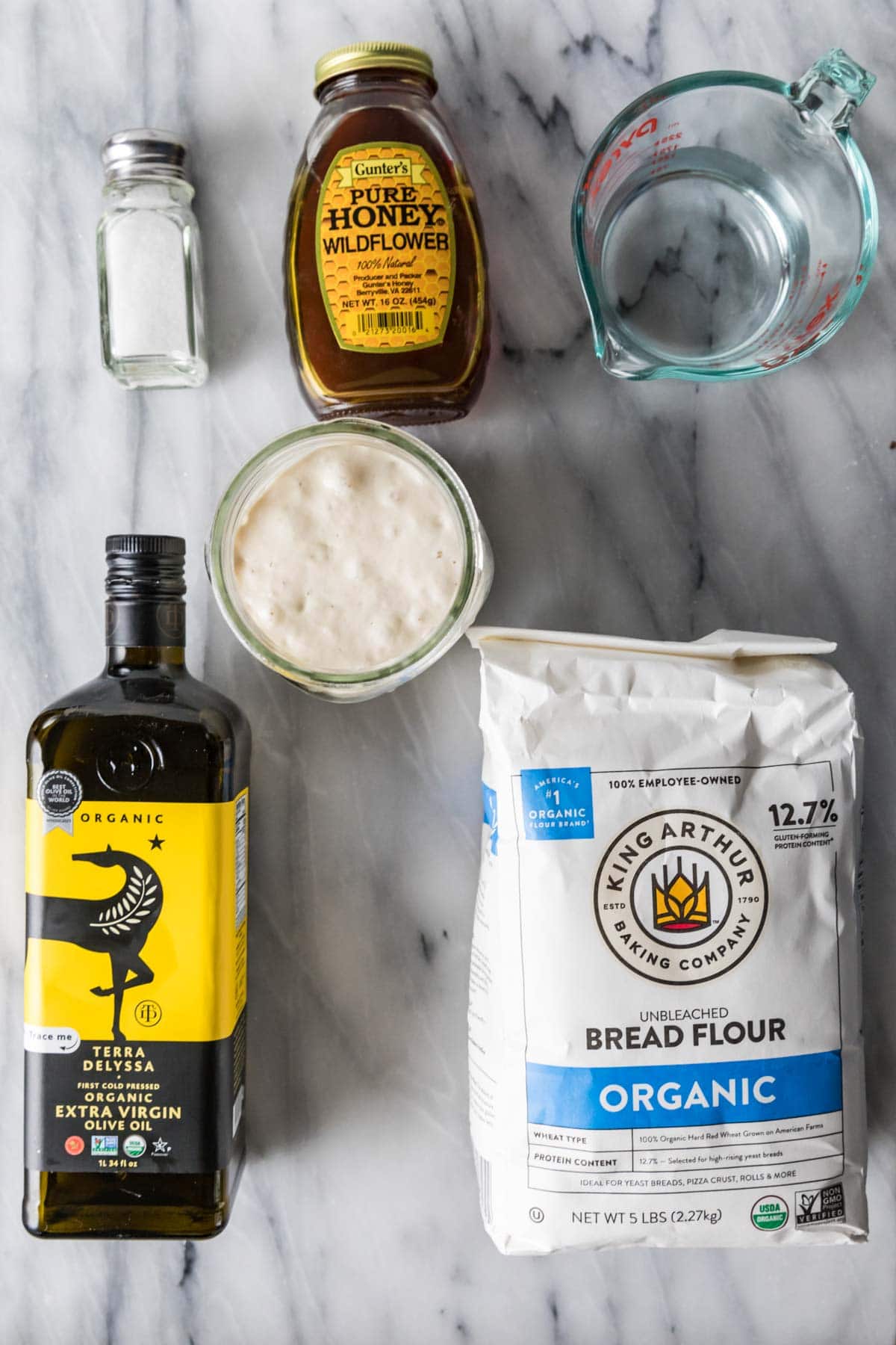 Overhead view of ingredients including bread flour, olive oil, honey, sourdough starter, and more.