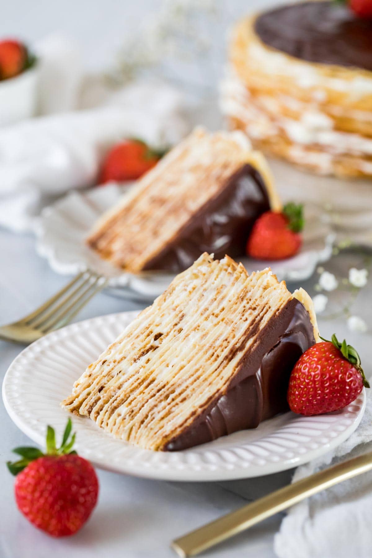 Slices of crepe cake topped with chocolate ganache on white plates.