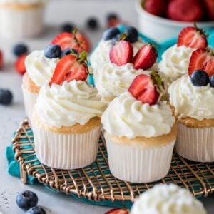 Angel food cupcakes topped with whipped cream and berries on a metal cooling rack.