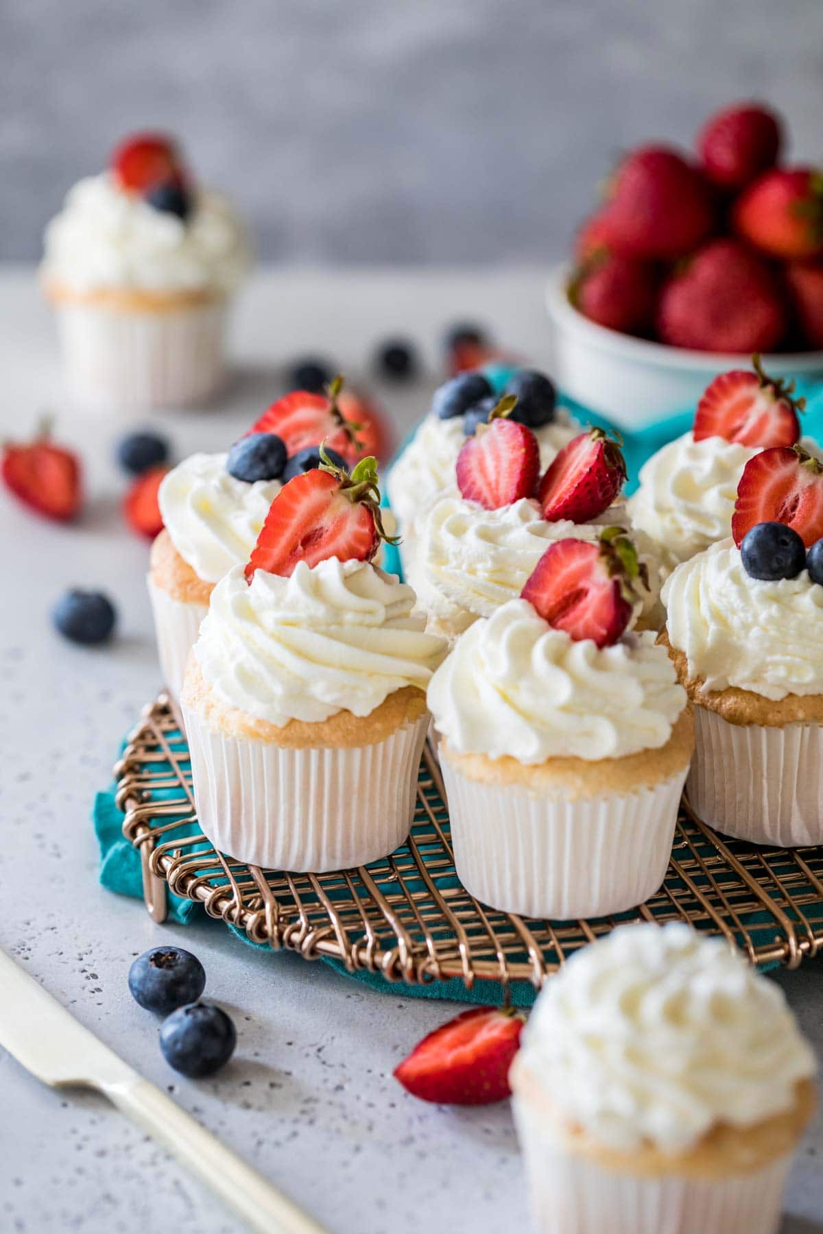 Angel food cupcakes topped with whipped cream and berries on a metal cooling rack.