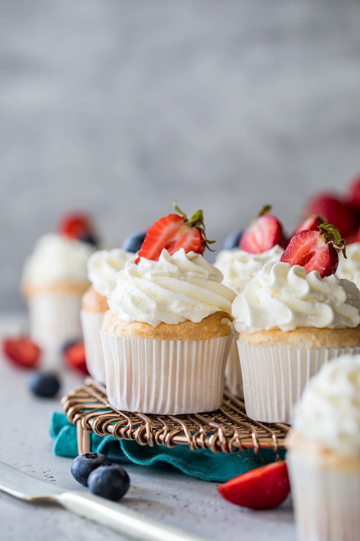 Cupcakes topped with piped whipped cream and strawberries on a metal cooling rack.