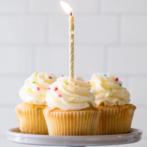 Four birthday cupcakes on a cake stand with a lit gold birthday candle in the front cupcake.