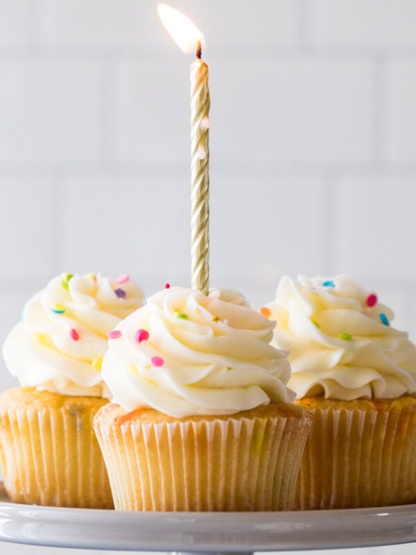 Four birthday cupcakes on a cake stand with a lit gold birthday candle in the front cupcake.