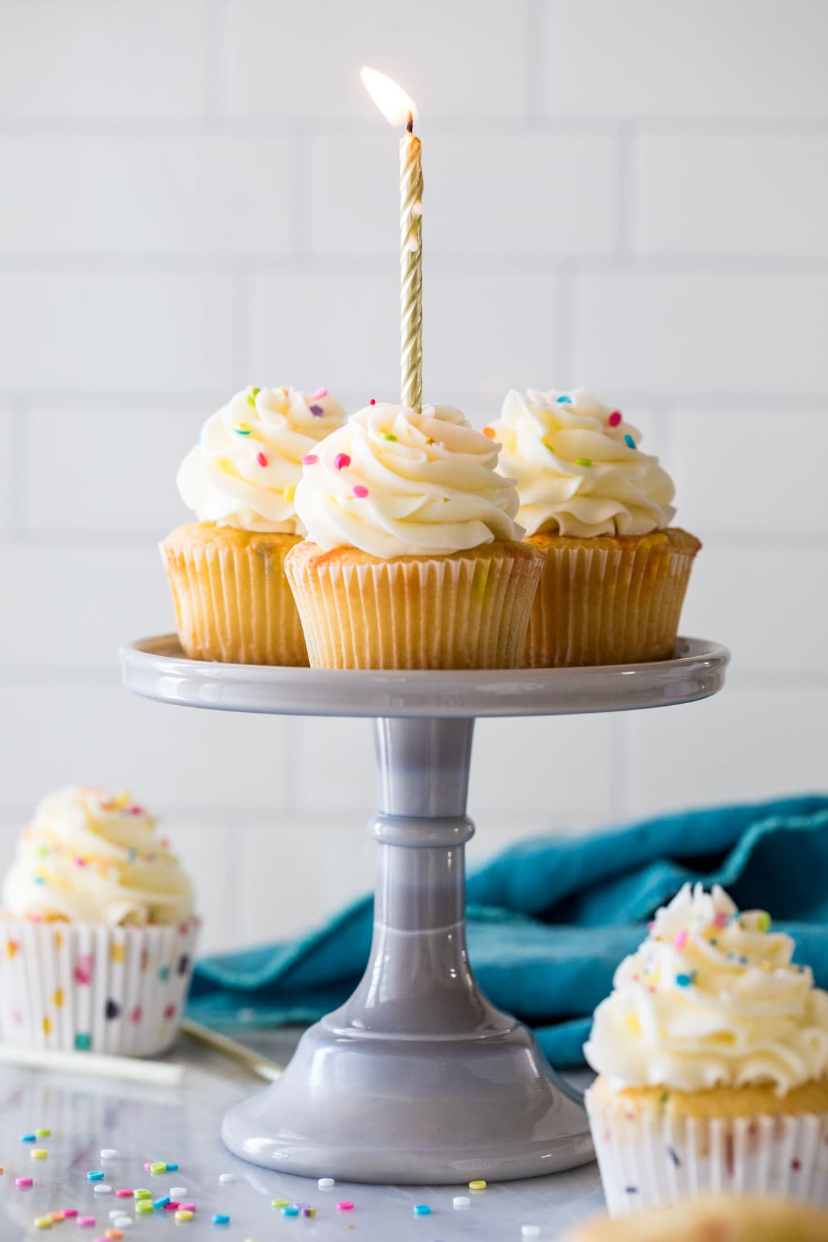 Four birthday cupcakes on a cake stand with a lit gold birthday candle in the front cupcake.