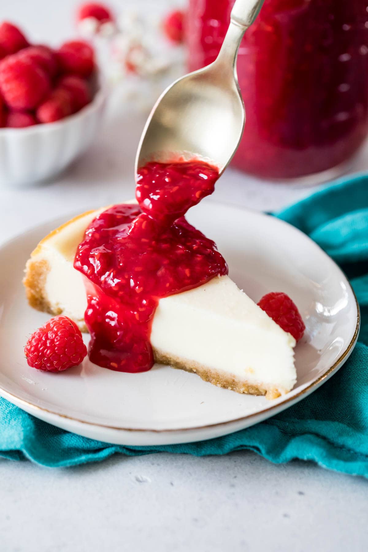 Spoon pouring raspberry sauce onto a slice of cheesecake.