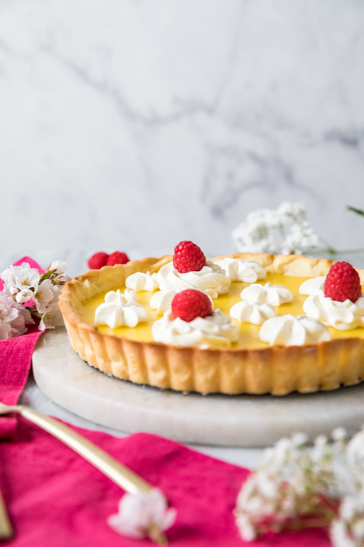 Head on view of a tart filled with lemon curd and garnished with whipped cream and raspberries.