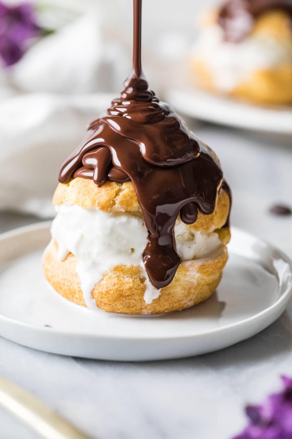 Chocolate ganache being poured on top of a choux bun filled with ice cream.