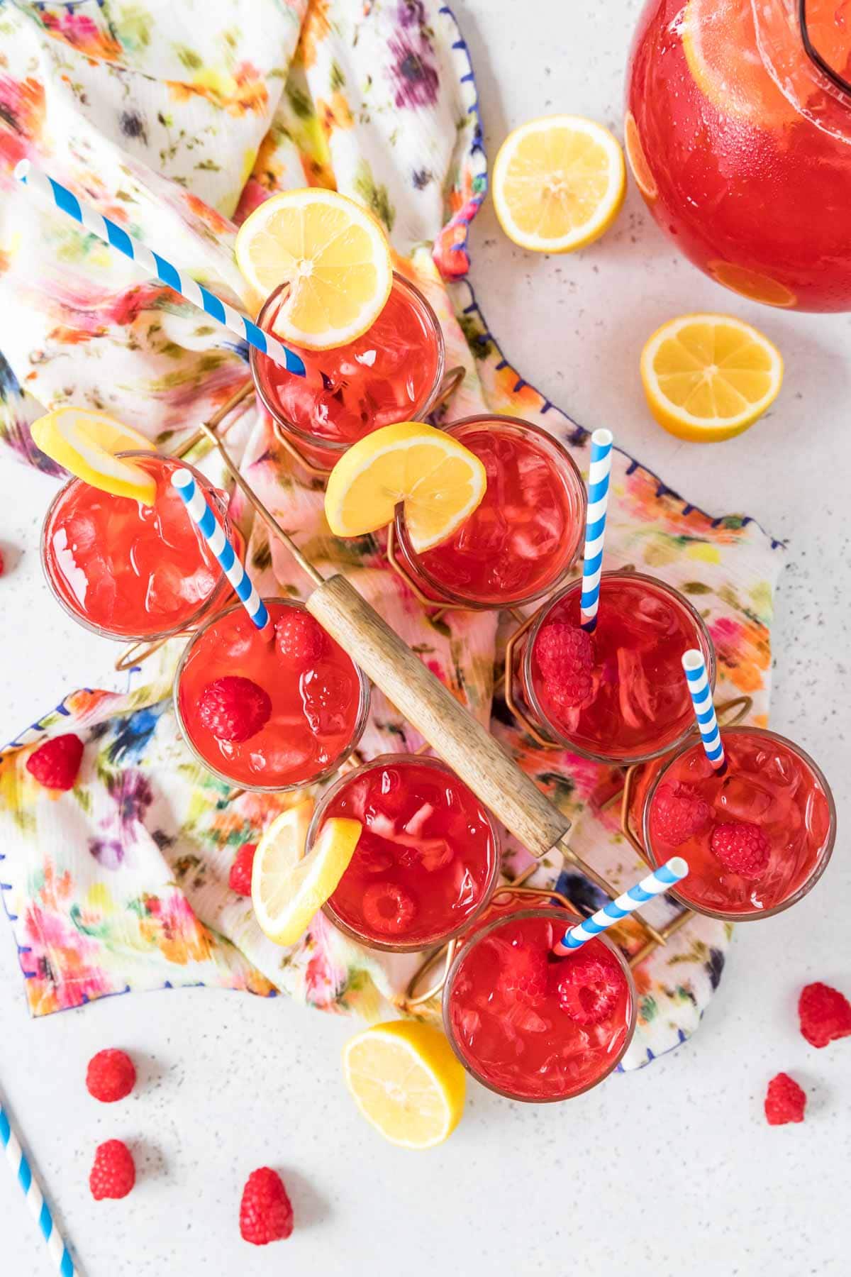 Overhead view of eight glasses of red raspberry lemonade garnished with lemon slices and topped with striped blue and white straws.