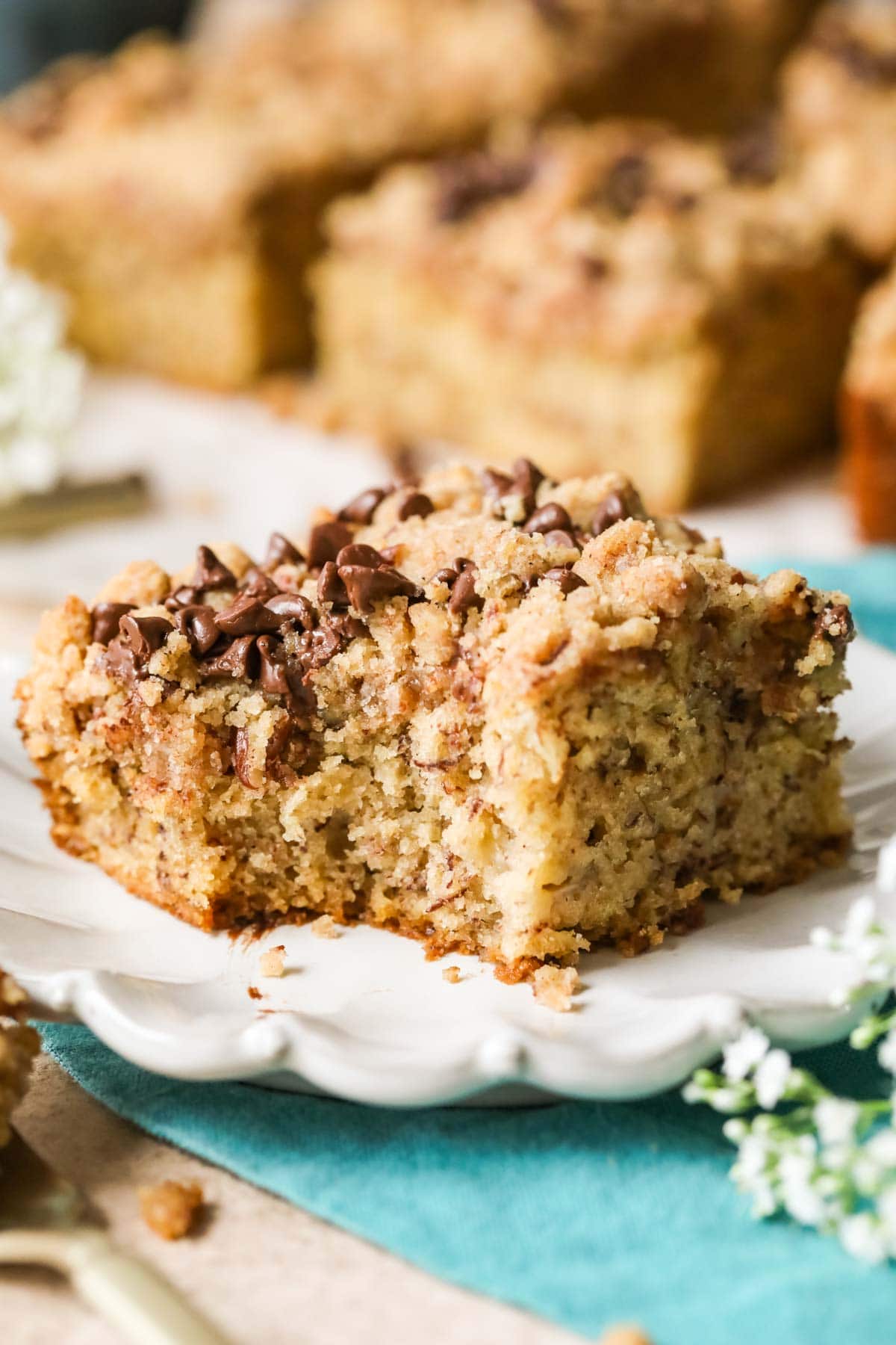Slice of streusel and chocolate chip topped banana coffee cake on a white plate with one bite missing.