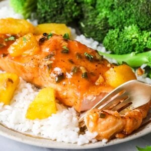 Fork cutting into maple glazed salmon served on a bed of rice with a side of broccoli.