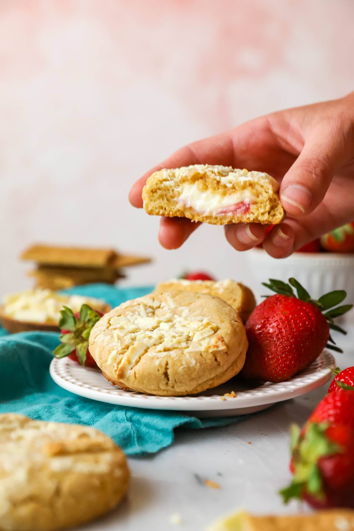 Hand pulling half of a strawberry cheesecake stuffed cookie off a plate of cookies.