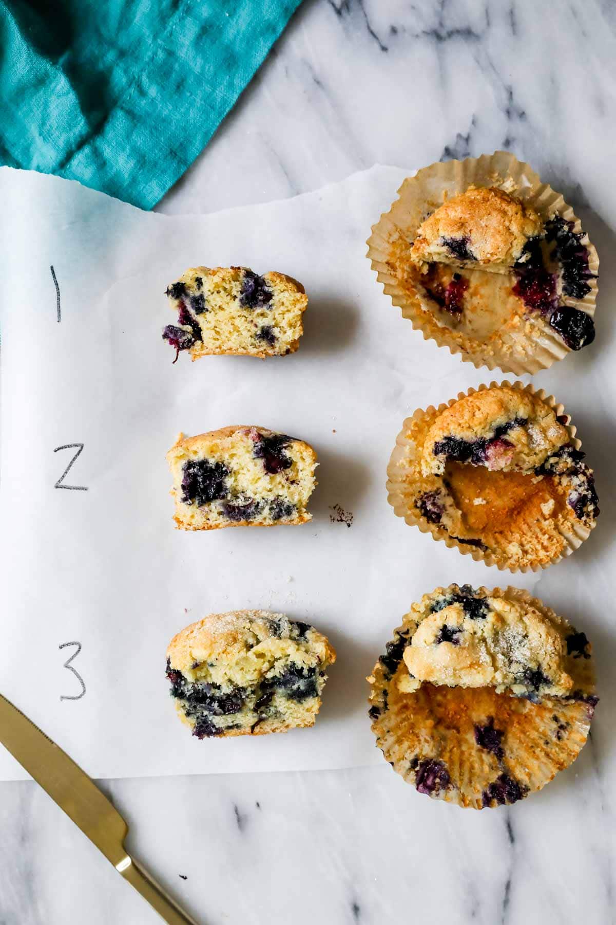 Overhead of 3 numbered blueberry muffins cut in half. 1 is shrunken with dense dry crumb, 2 is less shrunken with tight crumb, 3 has juicy berries and plush crumb 