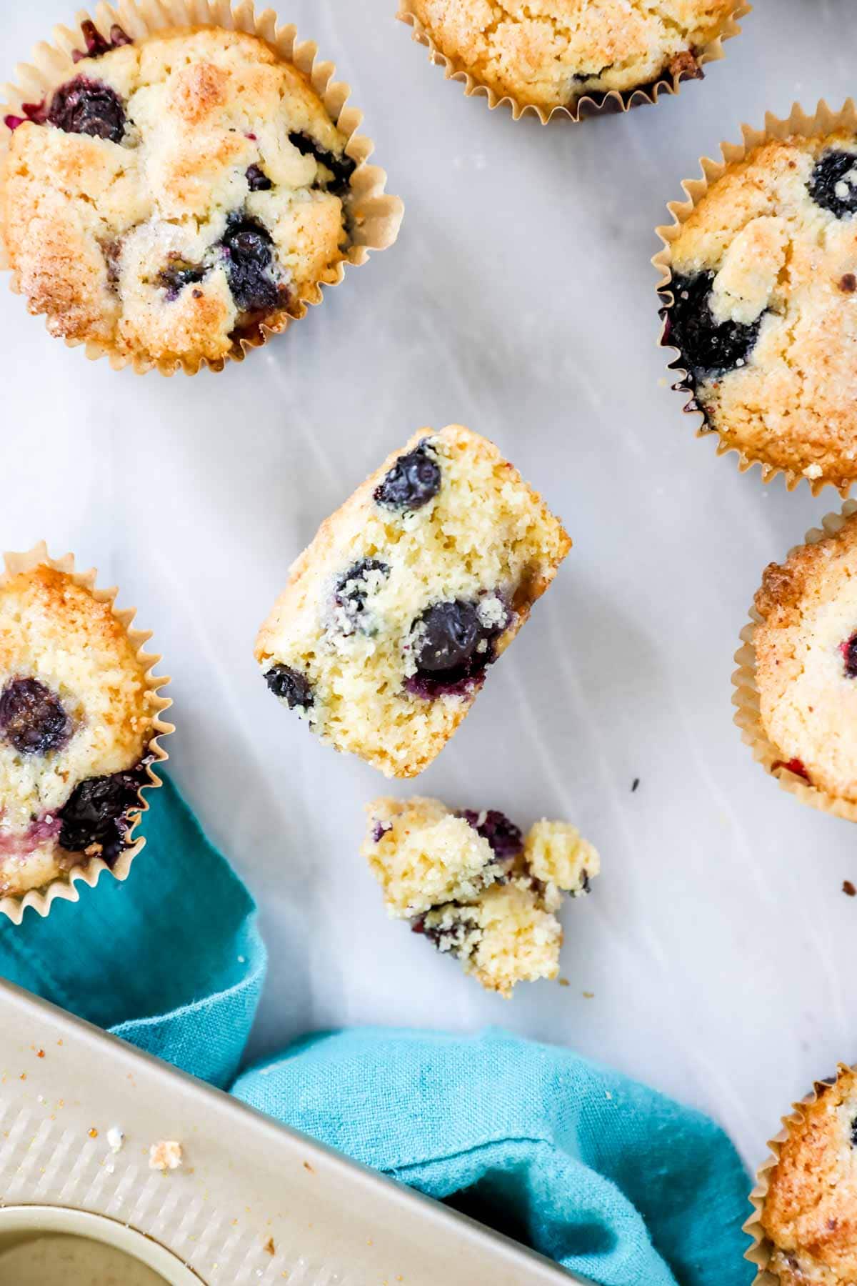 Overhead of blueberry muffin cut in half surrounded by intact muffins. The crumb appears dry and like cornbread