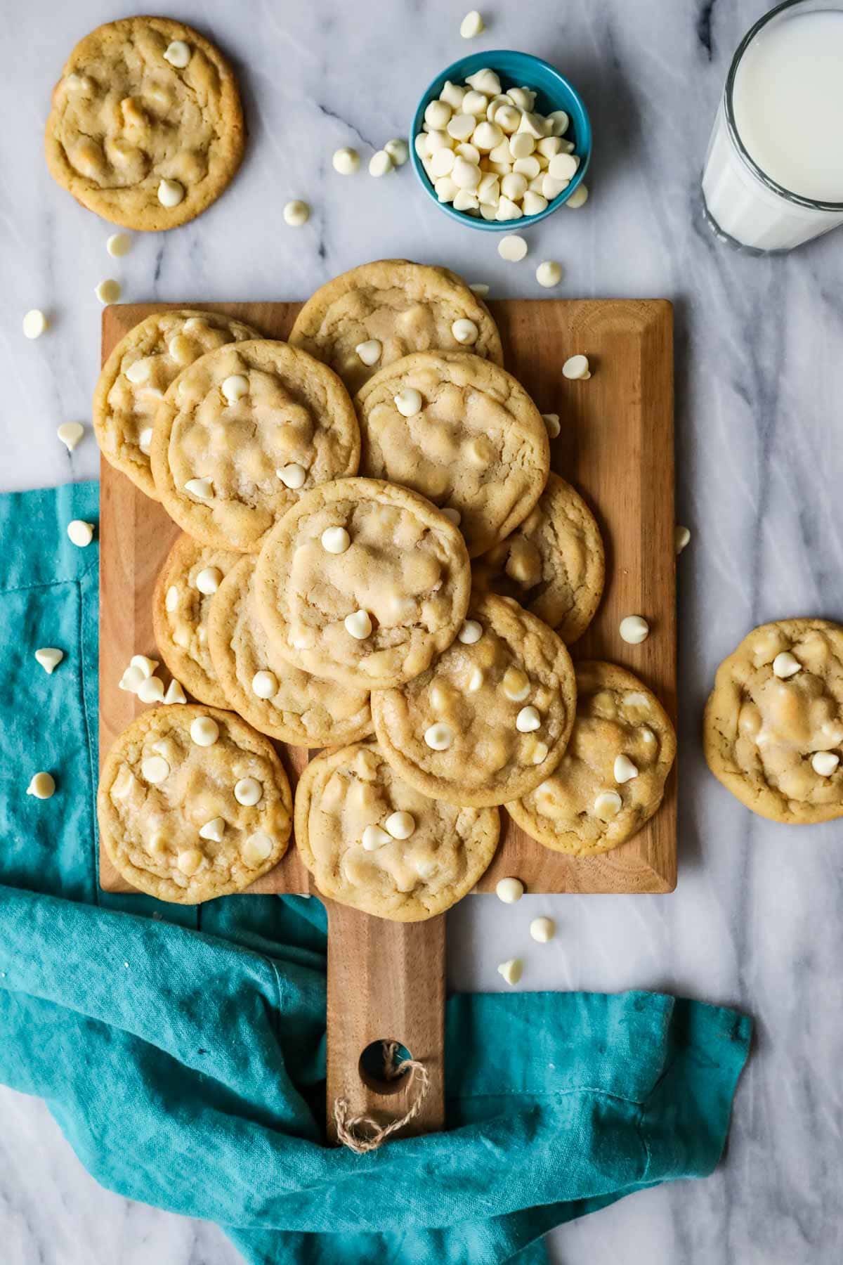 Overhead view of cookies studded with white chocolate chips scattered across a wood cutting board.