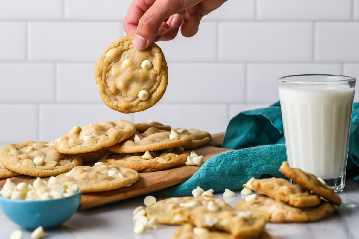Hand holding a white chocolate chip cookie above a pile of other cookies and a glass of milk.