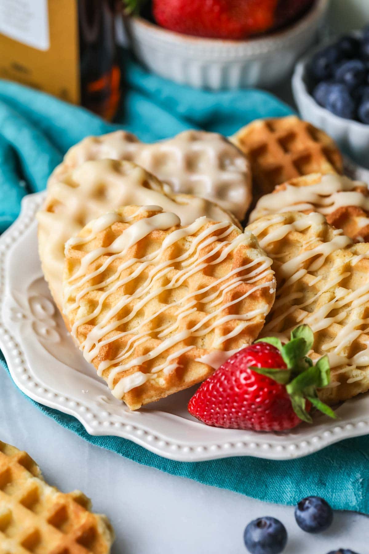 Heart shaped waffle cookies with a drizzled maple glaze on a white plate with a strawberry.