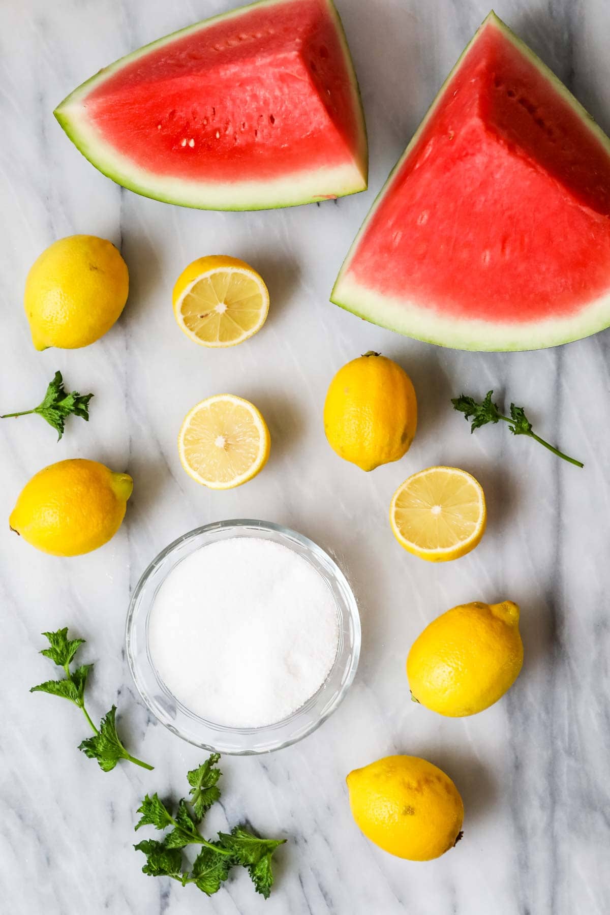 Overhead view of watermelon slices, lemon halves, mint leaves, and sugar.