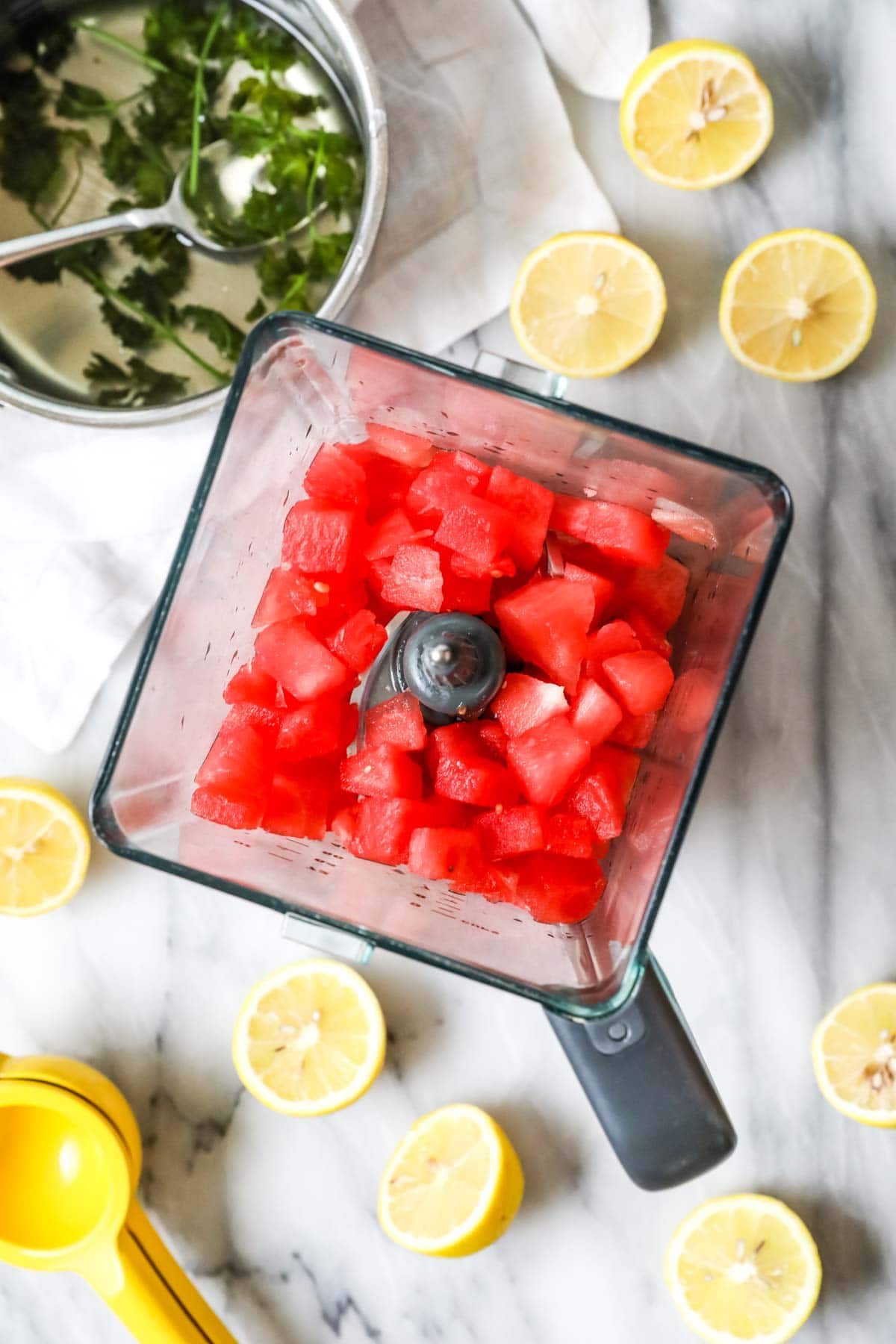 Overhead view of a blender full of watermelon chunks surrounded by lemon halves and a pot of mint simple syrup.