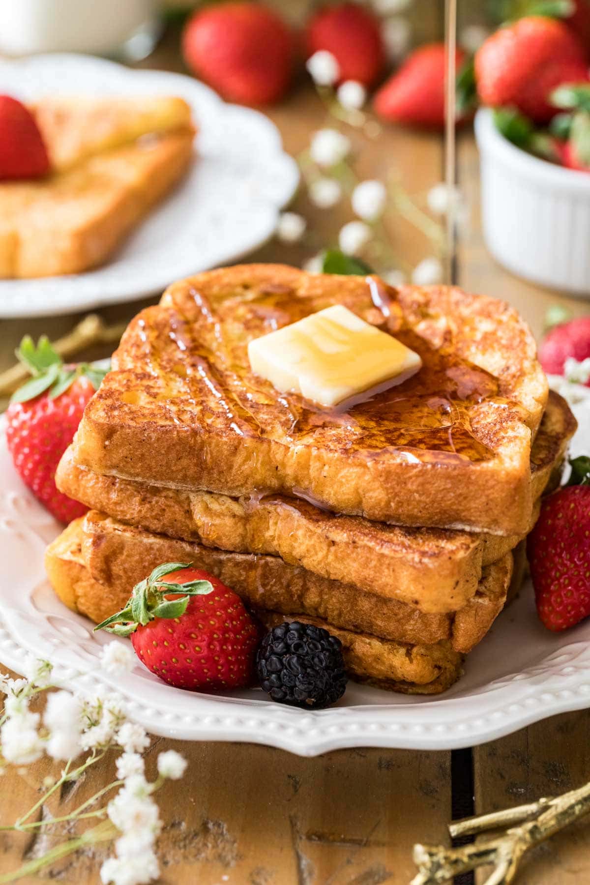 Syrup being drizzled over a stack of french toast with butter on top and berries scattered around.