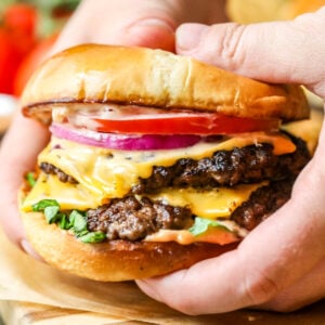 Hands holding a smash burger topped with American cheese, onion, tomato, and lettuce.