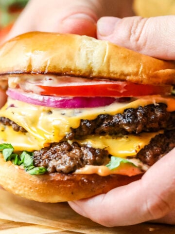 Hands holding a smash burger topped with American cheese, onion, tomato, and lettuce.