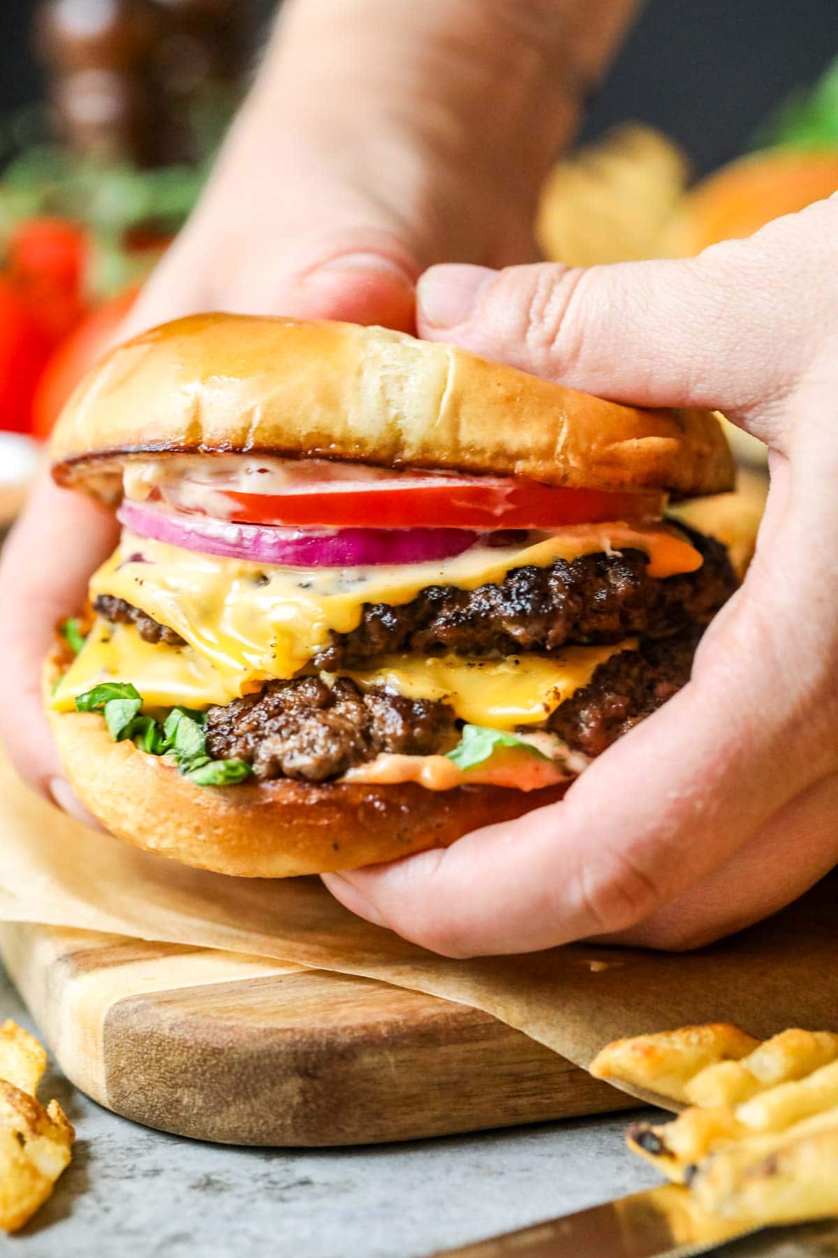Hands holding a smash burger topped with American cheese, onion, tomato, and lettuce.