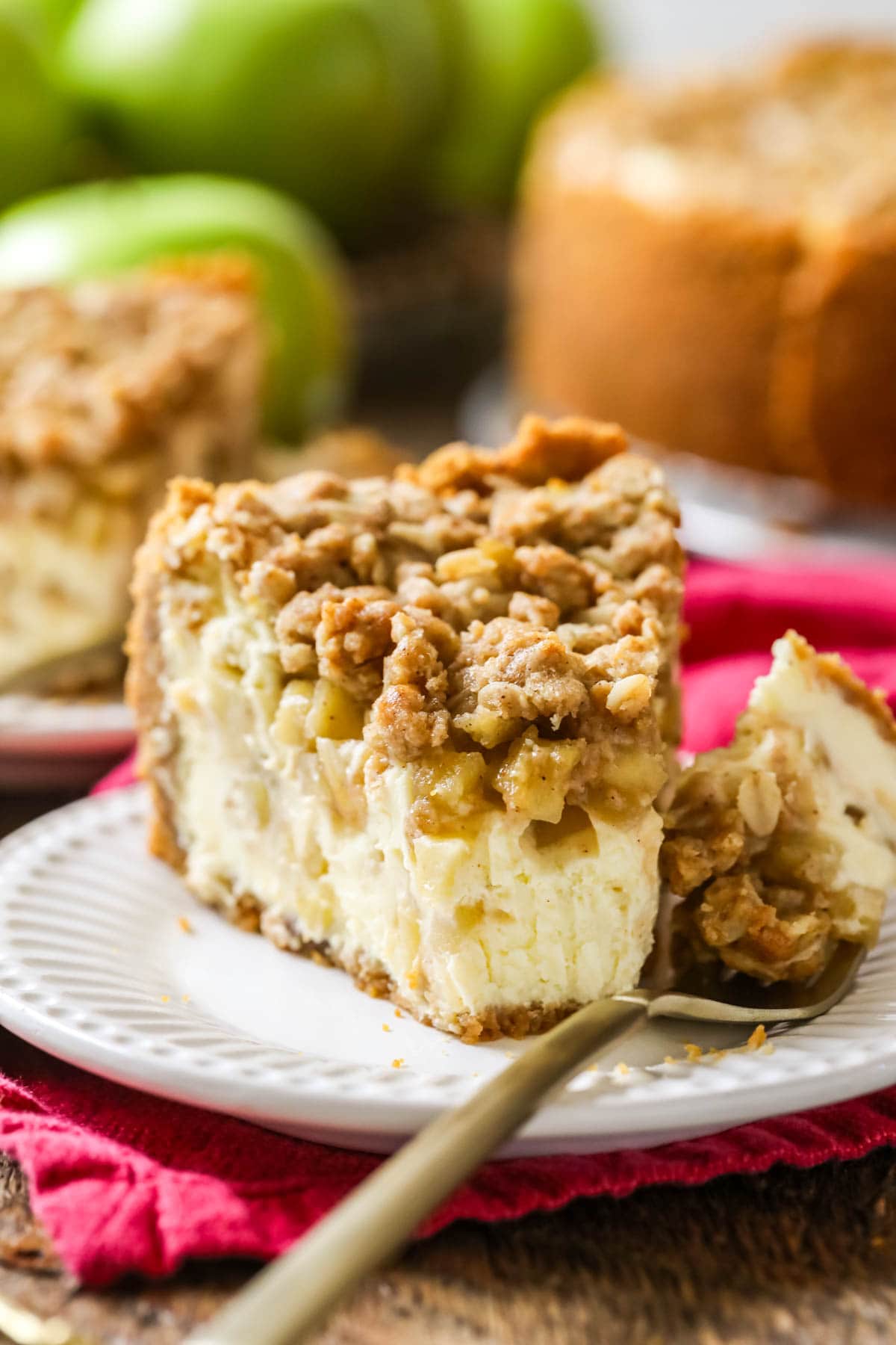 Slice of cheesecake made with apple pie filling and streusel topping on a plate with one bite sitting on a fork to the side.