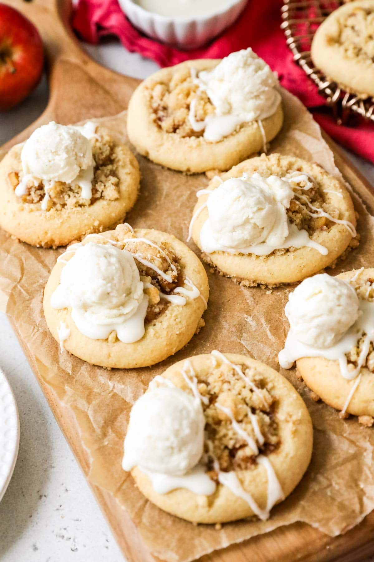 Overhead view of six cookies topped with baked apples, streusel, vanilla glaze, and a scoop of buttercream "ice cream" on a wood cutting board.