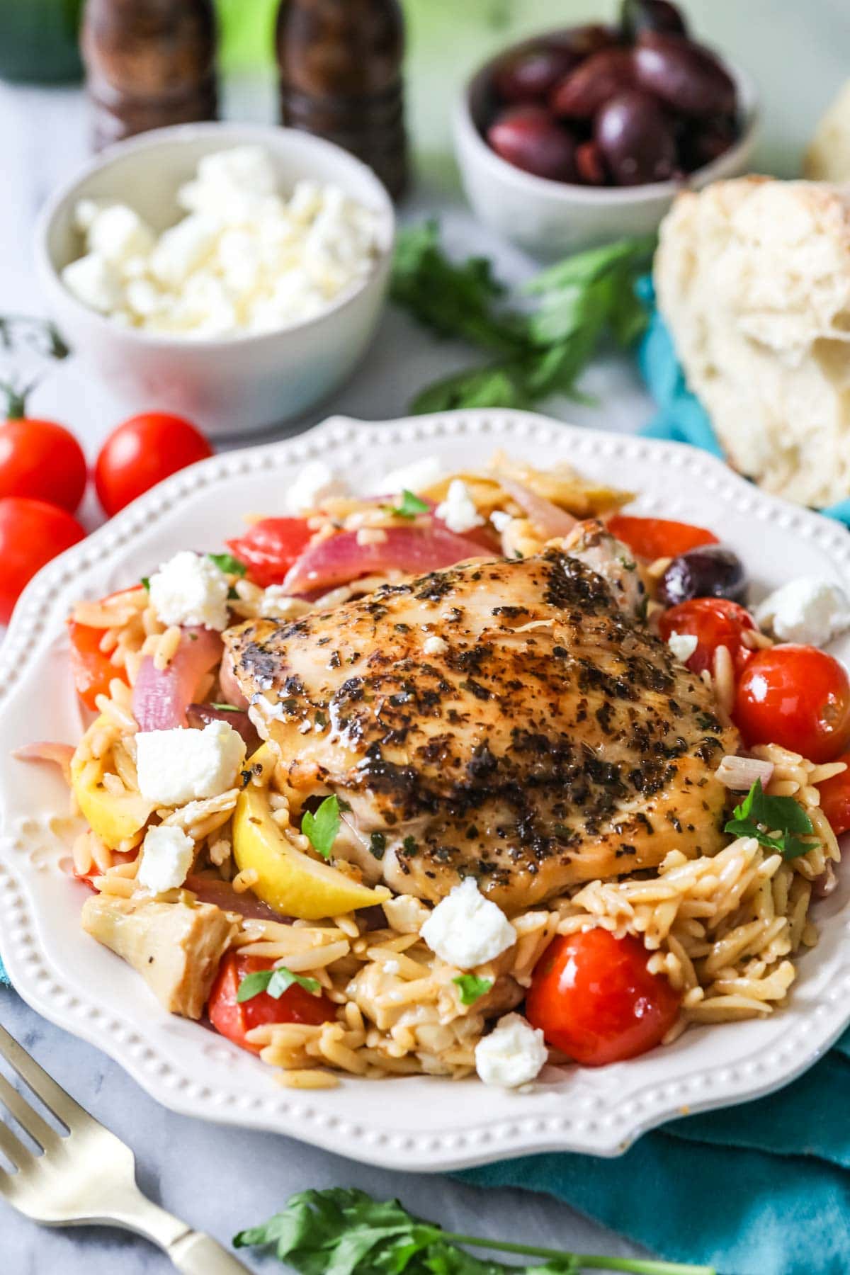 Plate of orzo topped with a seasoned chicken thigh, tomatoes, feta, lemon wedges, and more.