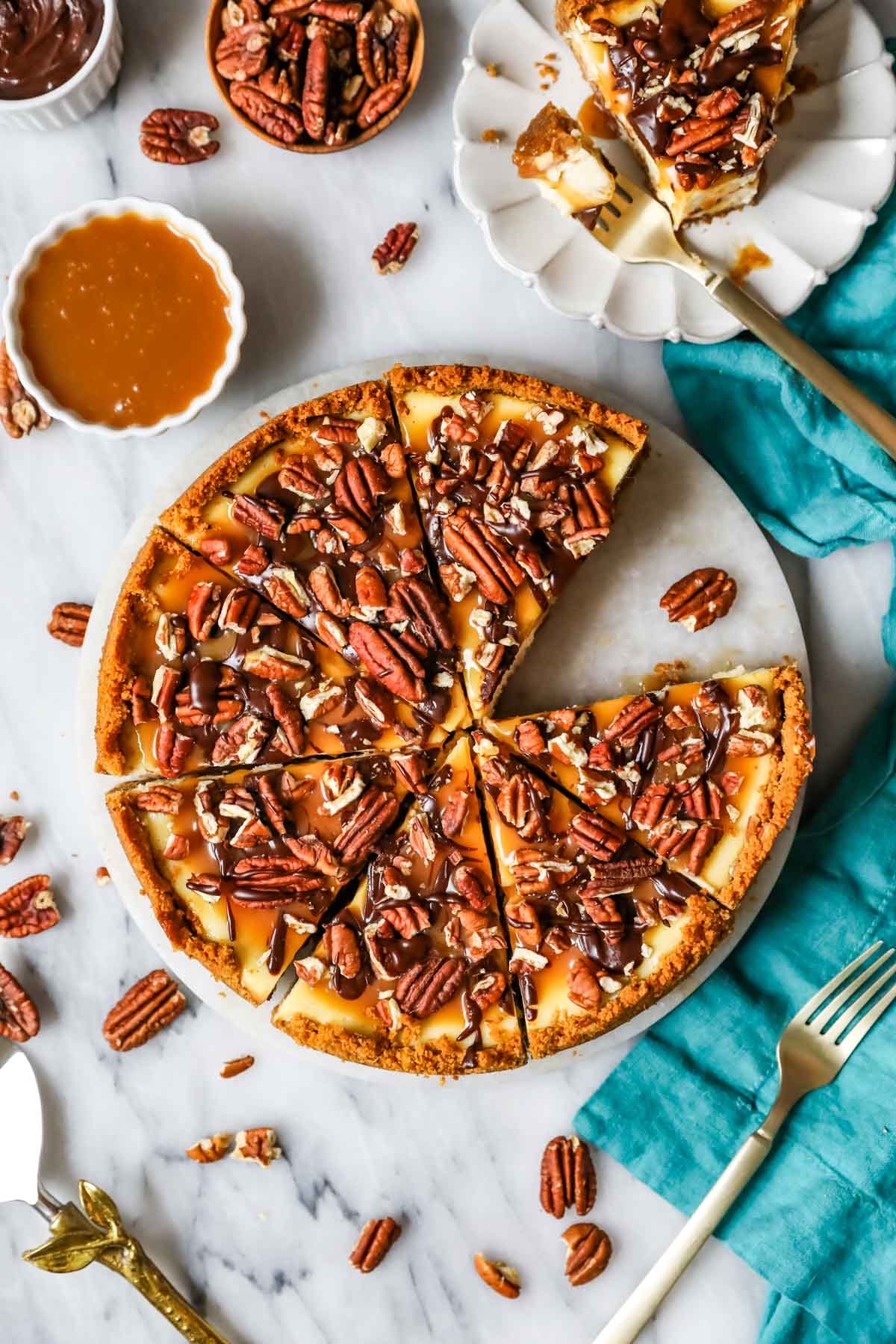 Overhead view of a cheesecake topped with caramel, pecans, and chocolate that's been cut into slices with one slice removed.