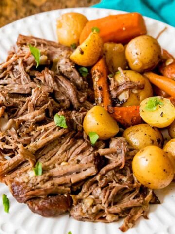 Juicy pot roast shredded on a plate beside roasted carrots and potatoes.