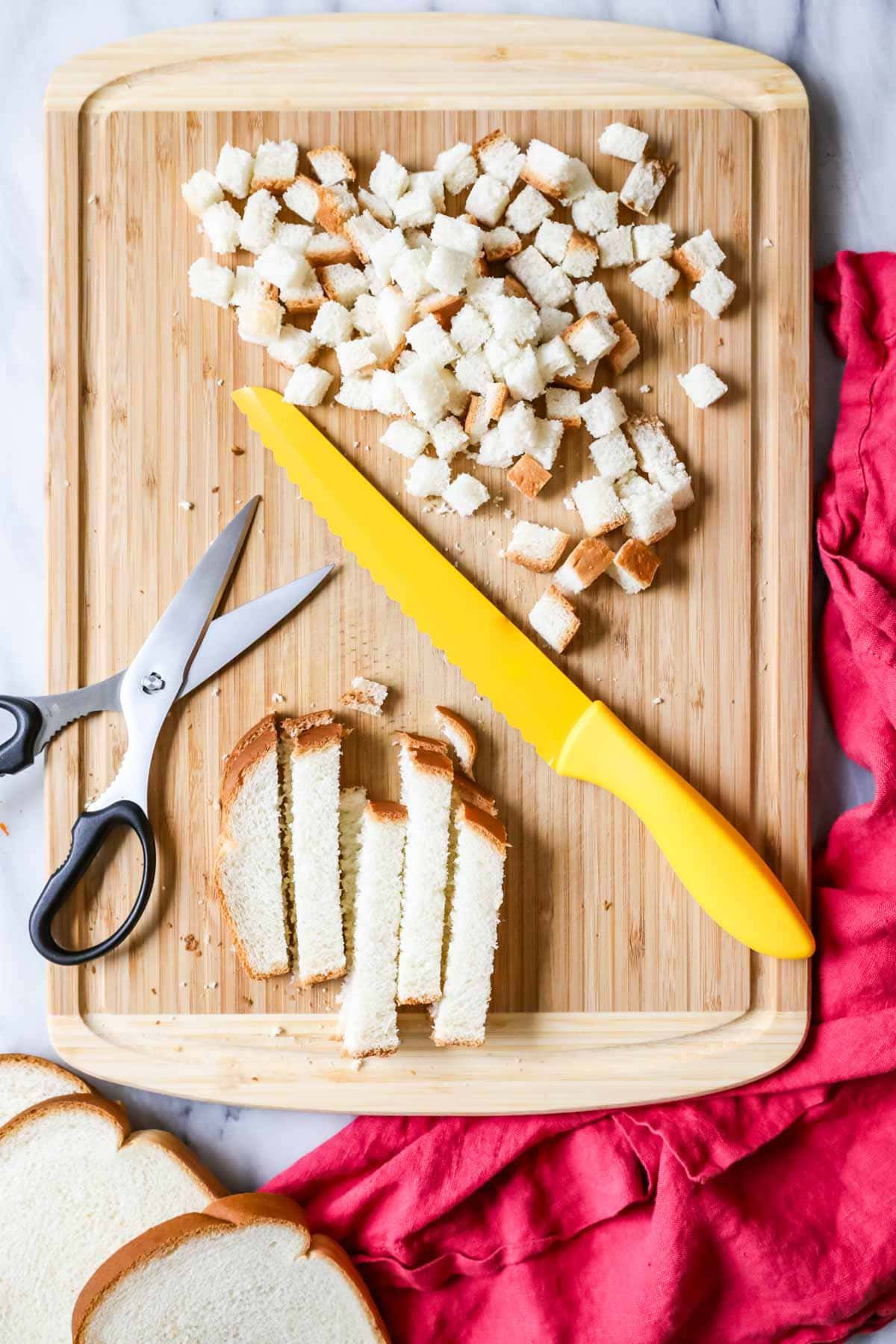 Overhead view of bread slices being cut into cubes on a cutting board.