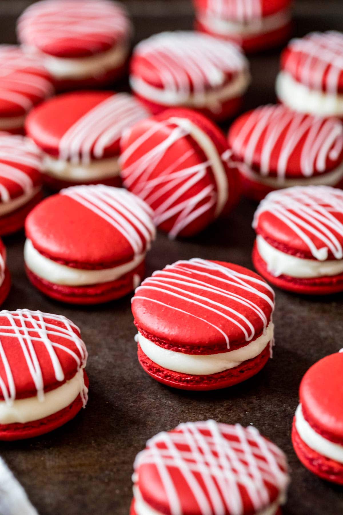 Red velvet macarons with a thin white chocolate drizzle on top resting on a dark surface.