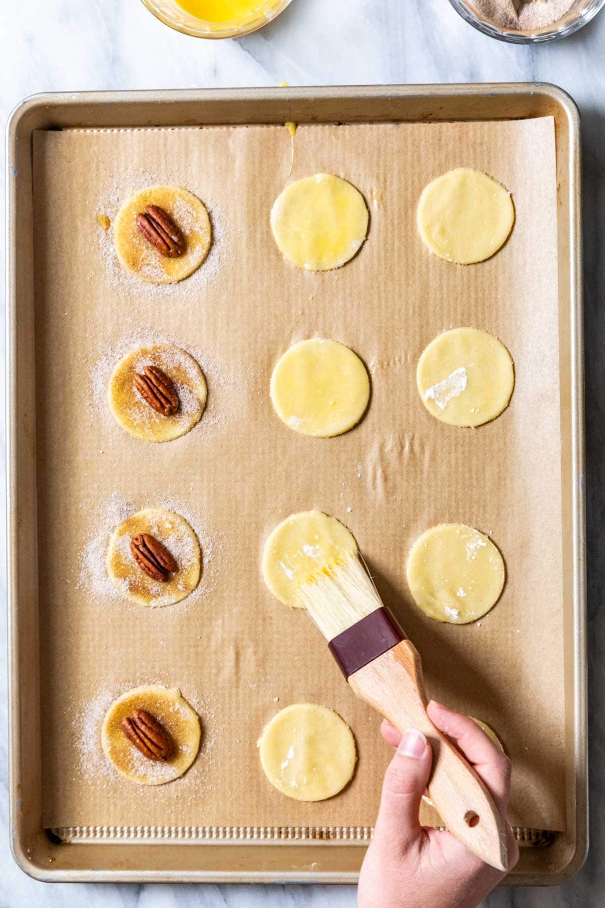 Overhead view of round cookie dough circles being brushed with egg wash, sprinkled with cinnamon sugar, and topped with pecan halves.