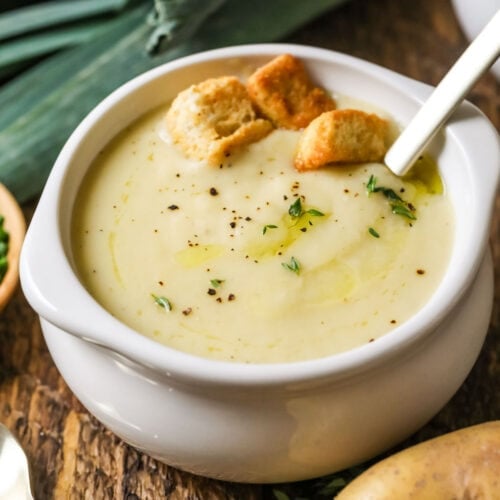 Small white crock of potato leek soup topped with croutons and chives.