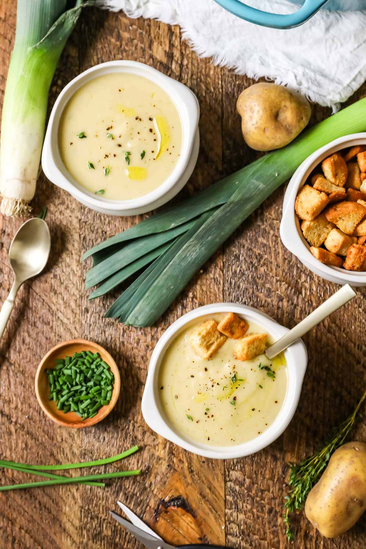 Overhead view of crocks of potato leek soup with leeks, chives, croutons, and potatoes surrounding them.
