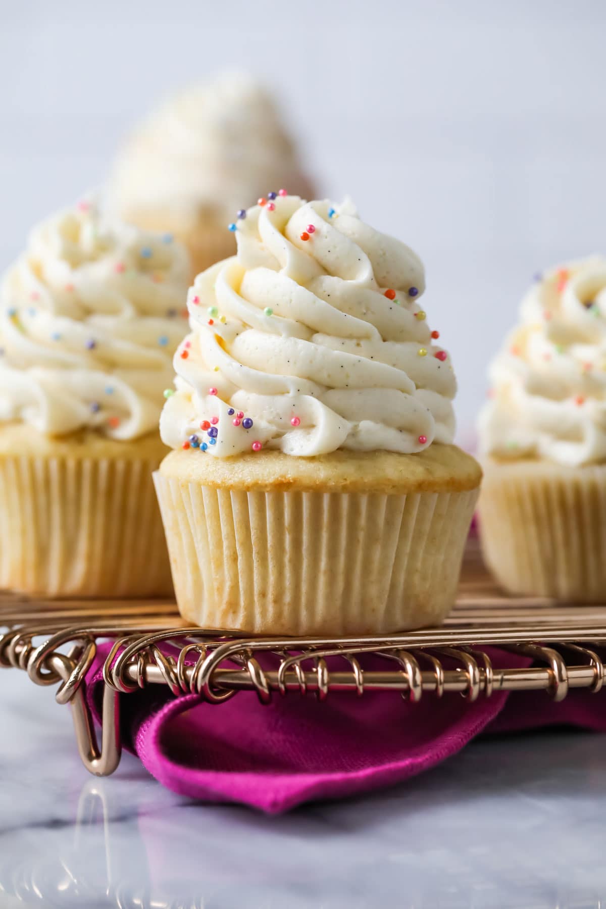 Vanilla cupcakes topped with tall swirls of vanilla bean buttercream on a cooling rack.