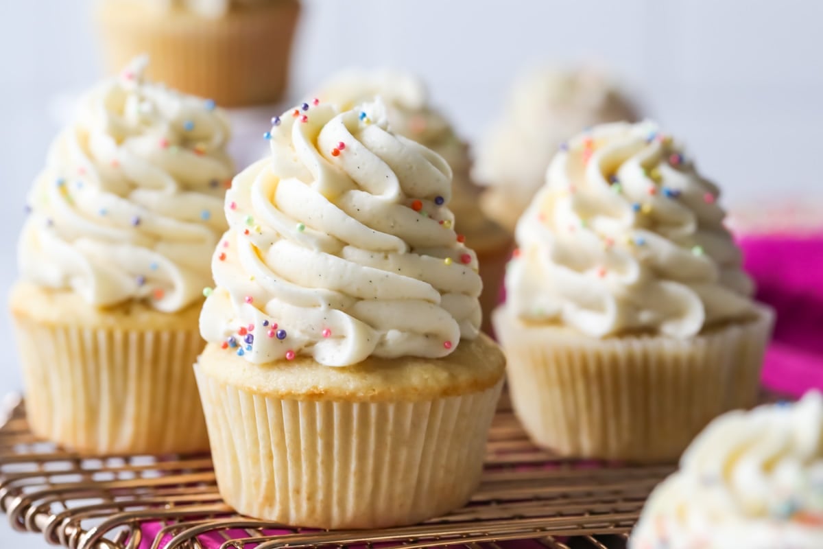 Cupcakes topped with tall piped swirls of frosting and rainbow sprinkles on a cooling rack.