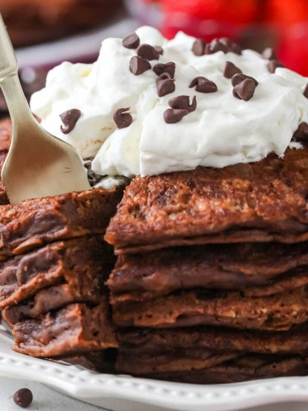 Fork taking a bite out of a stack of chocolate pancakes topped with whipped cream and chocolate chips.