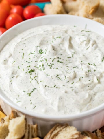 Bowl of dill dip with crackers, tomatoes, and bread surrounding it.