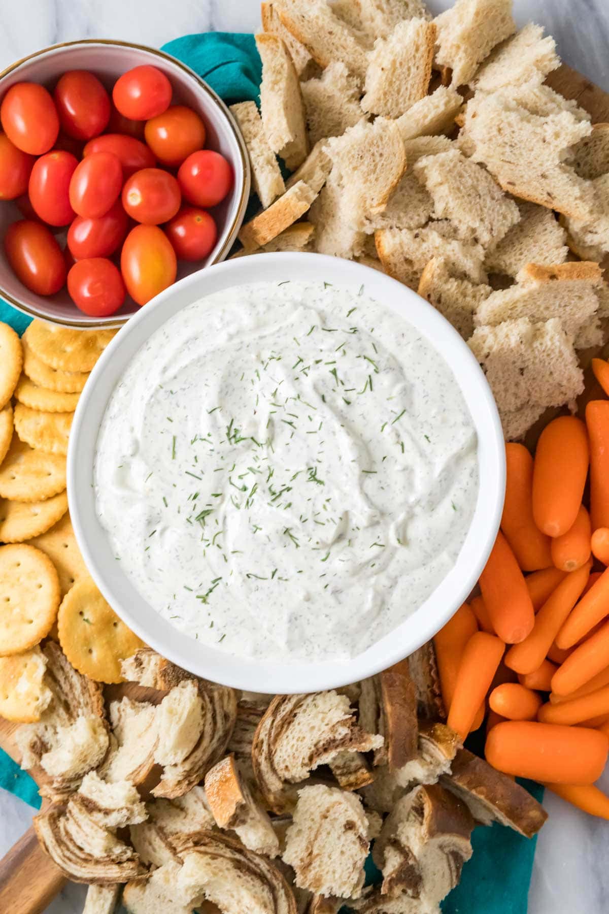 Overhead view of a bowl of dip surrounded by cherry tomatoes, celery, crackers, and torn bread pieces.