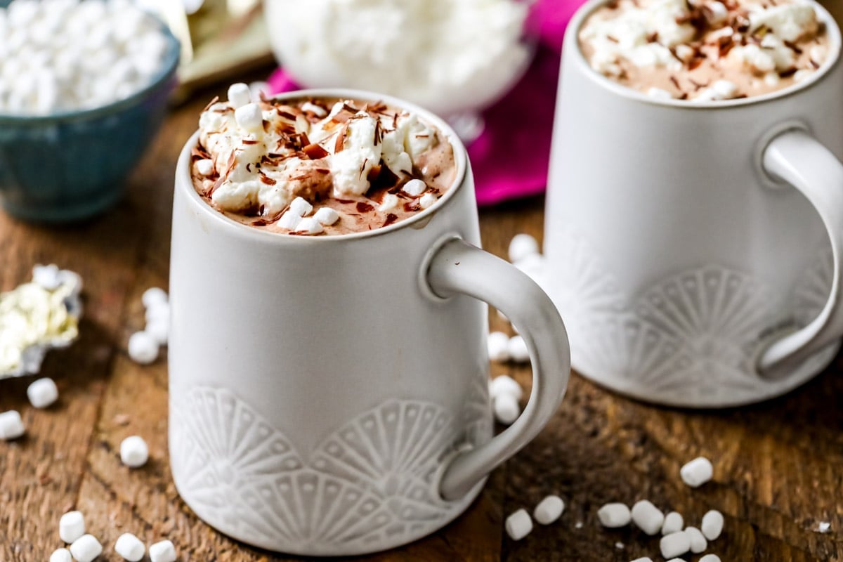 Two cups of cocoa topped with whipped cream, marshmallow, and chocolate shavings.